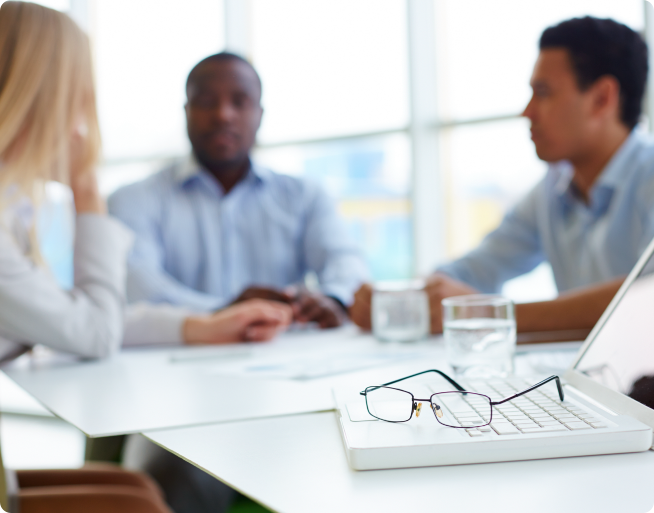 Glasses sit on a laptop computer as coworkers talk in the background