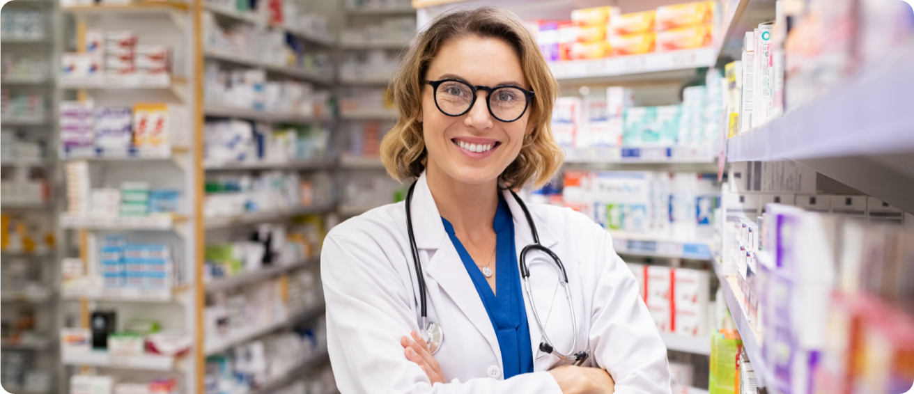 A smiling pharmacist standing in front of a wall full of medications.