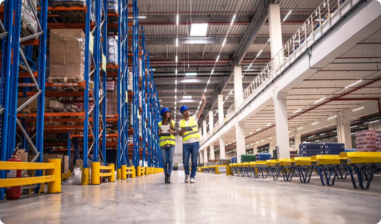 Two factory workers, wearing safety vests and hats, walking through a factory.
