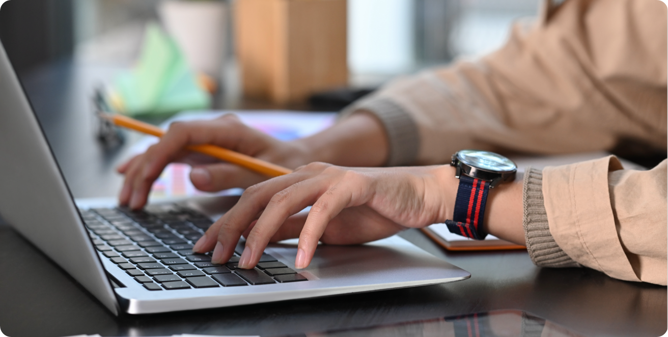 A person typing on a laptop computer while holding a pencil.