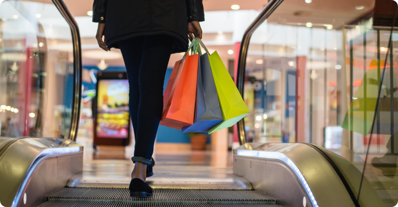 A person stepping of an escalator, holding three shopping bags.